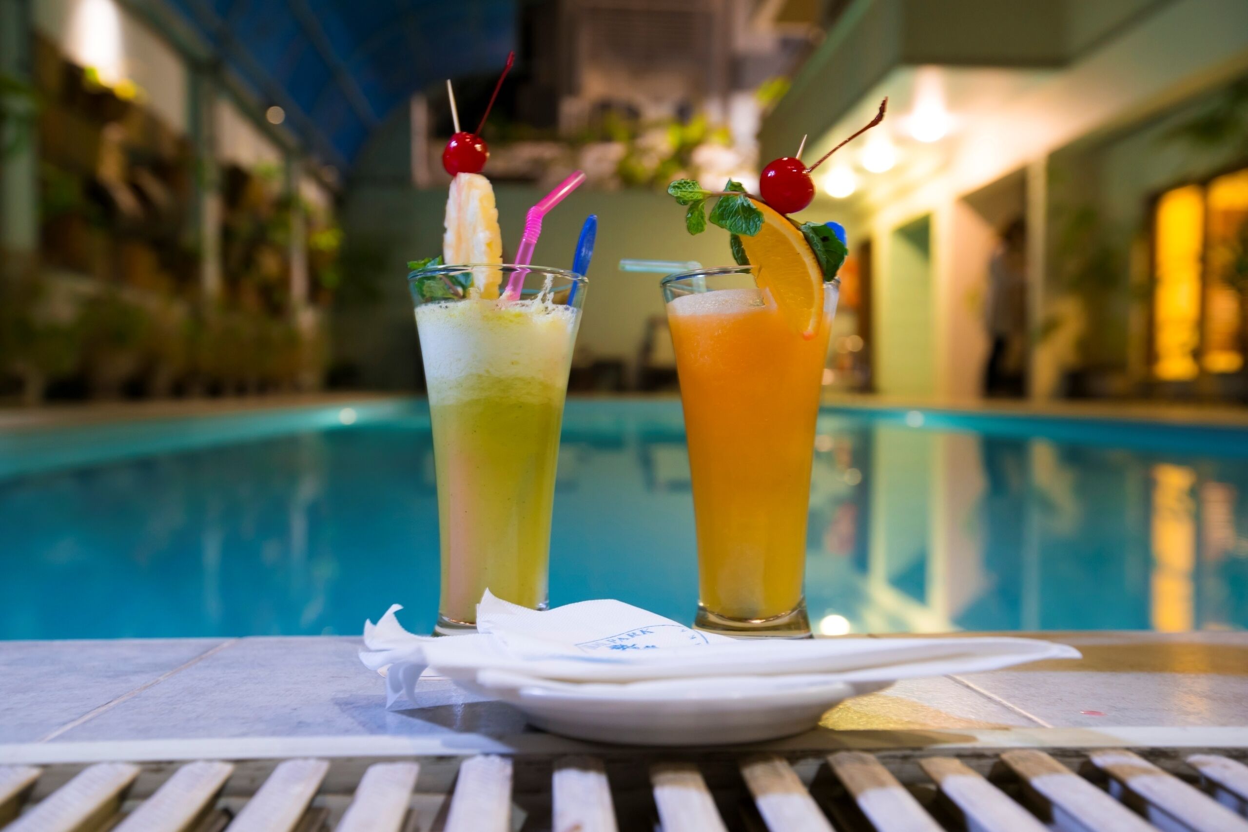 Two cocktails with fruit garnishes and straws on a table by a pool.
