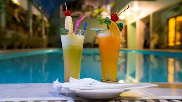 Two cocktails with fruit garnishes and straws on a table by a pool.