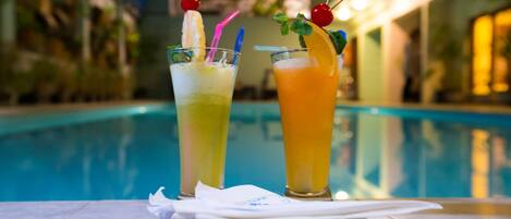 Dos cócteles con guarniciones de frutas y pajitas sobre una mesa junto a una piscina.