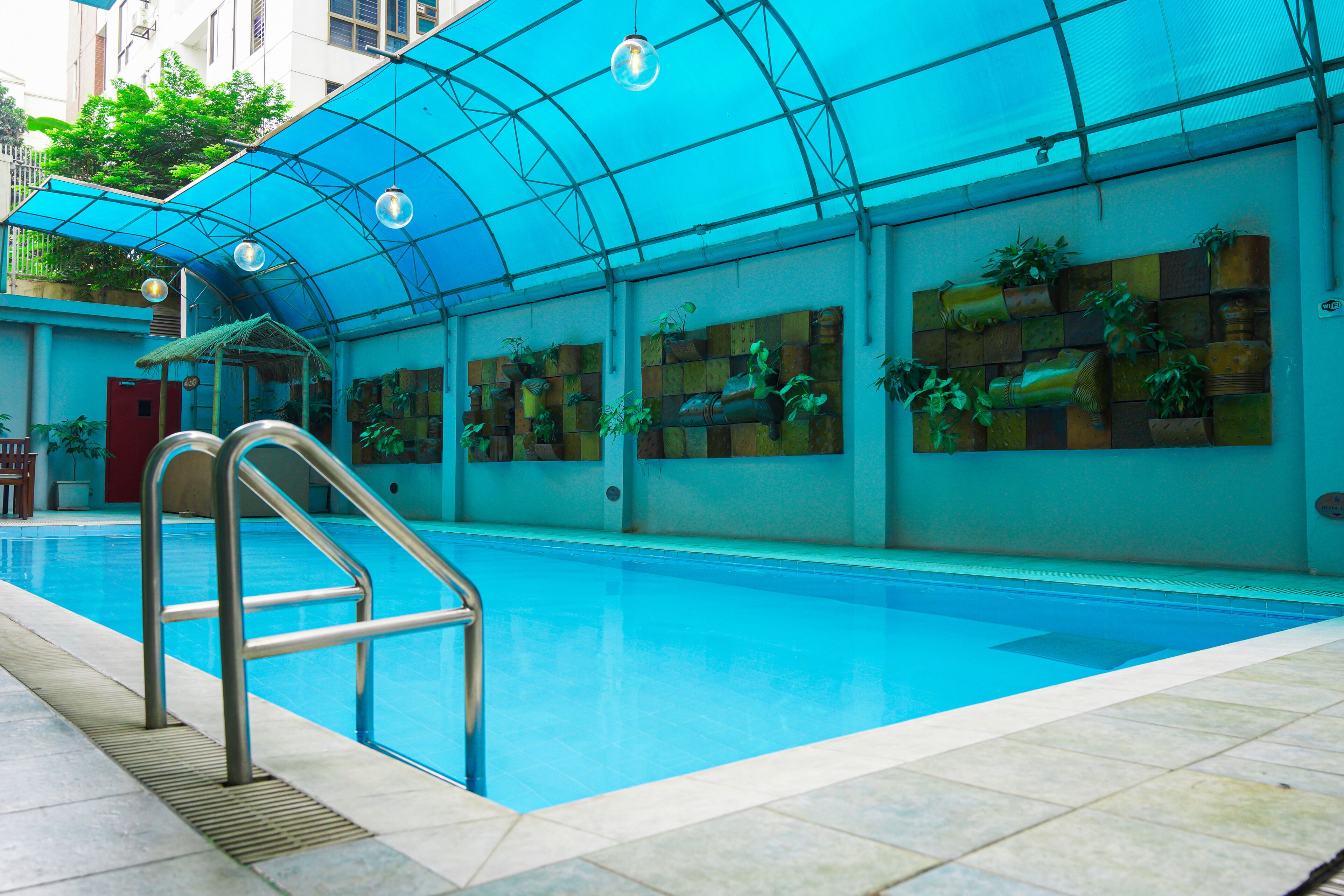 An indoor swimming pool with a blue-tiled floor, metal pool ladder, and a ceiling with a blue canopy.