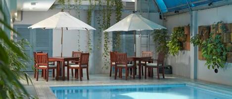 Un área de piscina con dos mesas y sillas de madera, rodeada de sombrillas, y una zona cubierta con plantas para sentarse.
