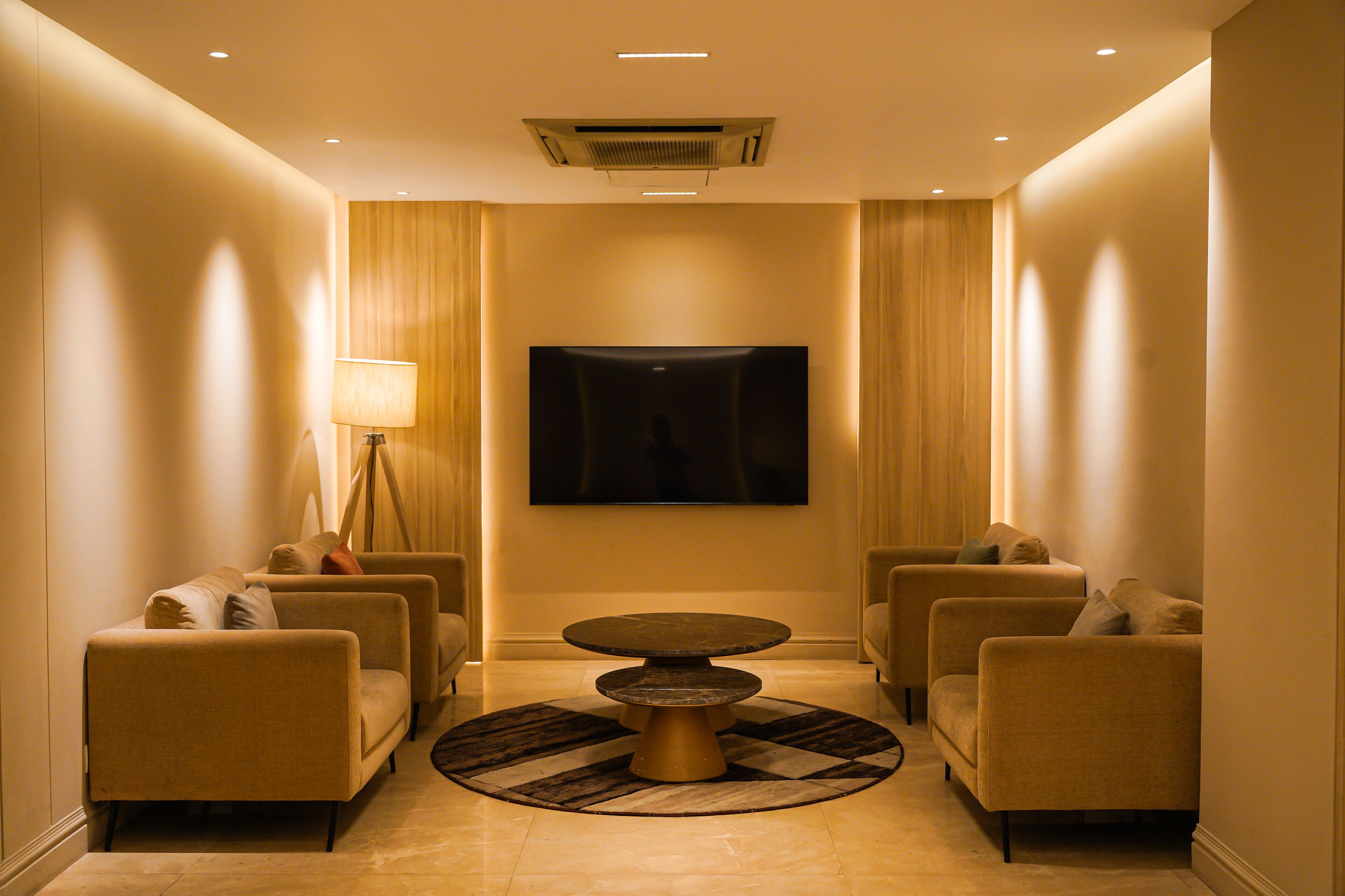 A hotel lobby with a round table, four sofas, a floor lamp, and a flat-screen TV on the wall.