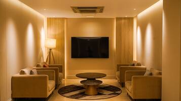 A hotel lobby with a round table, four sofas, a floor lamp, and a flat-screen TV on the wall.
