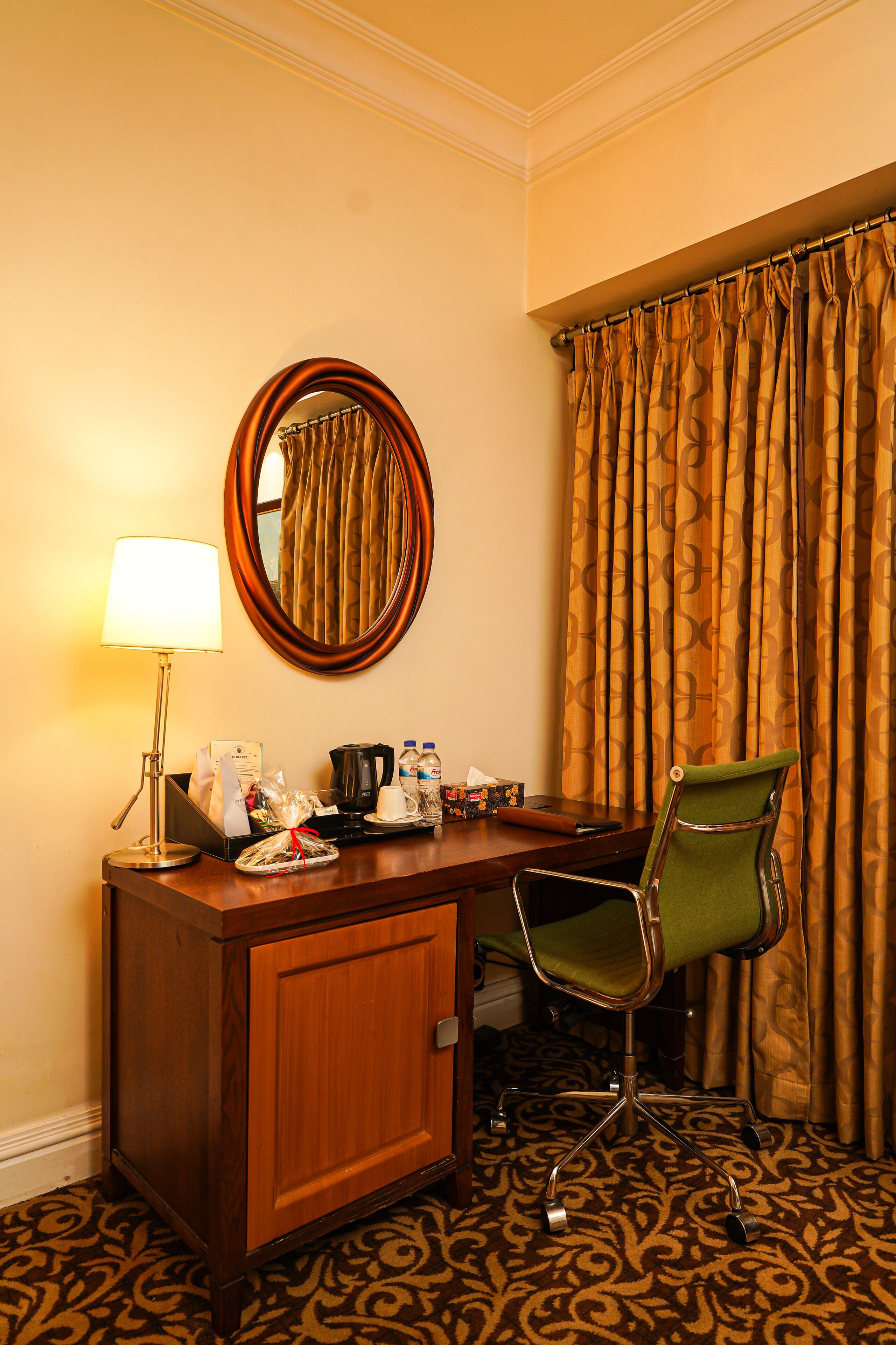 A hotel room with a wooden desk, a green chair, a lamp, a mirror, and a patterned carpet.