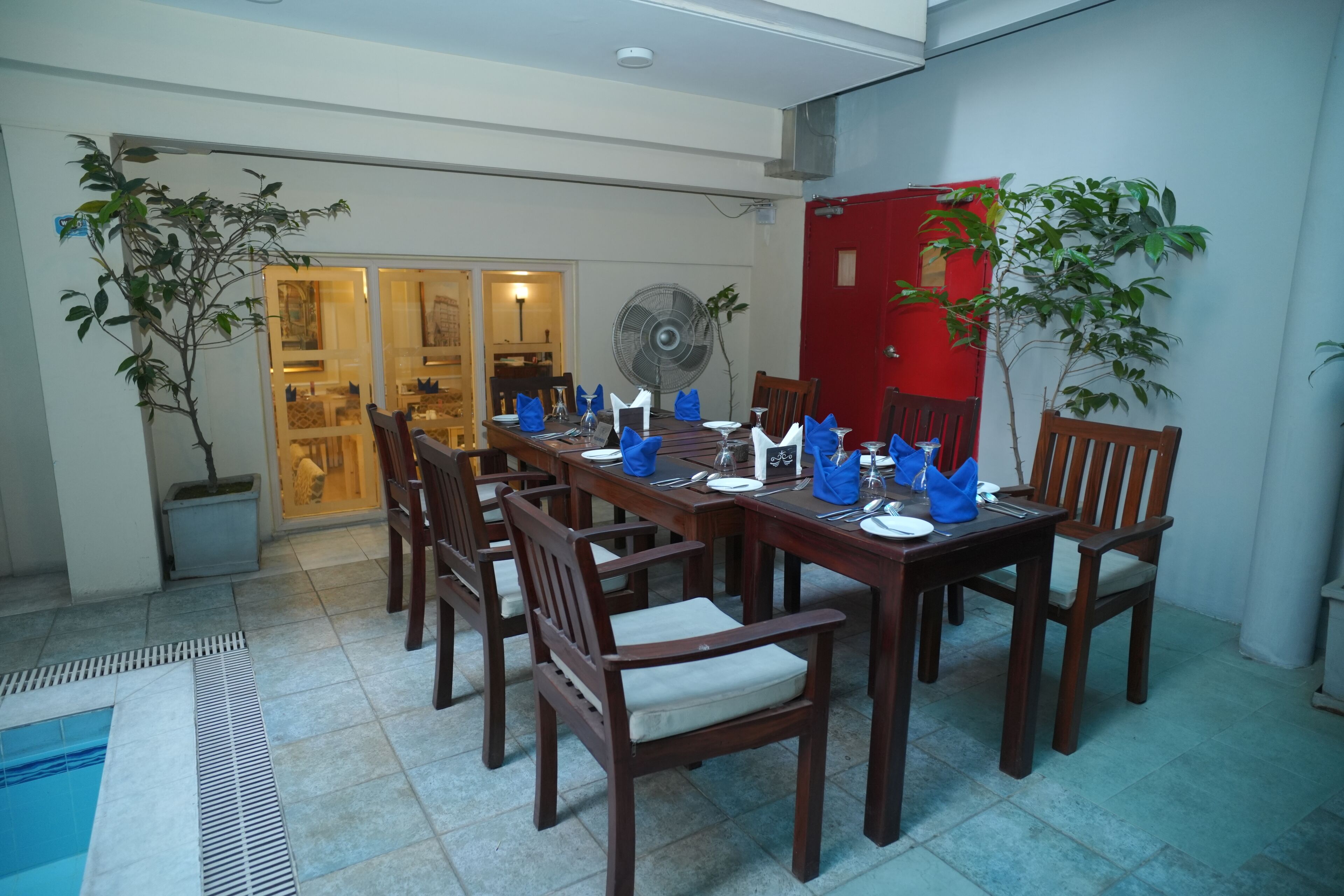 A dining area with a long wooden table set for six, surrounded by wooden chairs. There is a fan on the wall, and potted plants are placed on either side of the table.