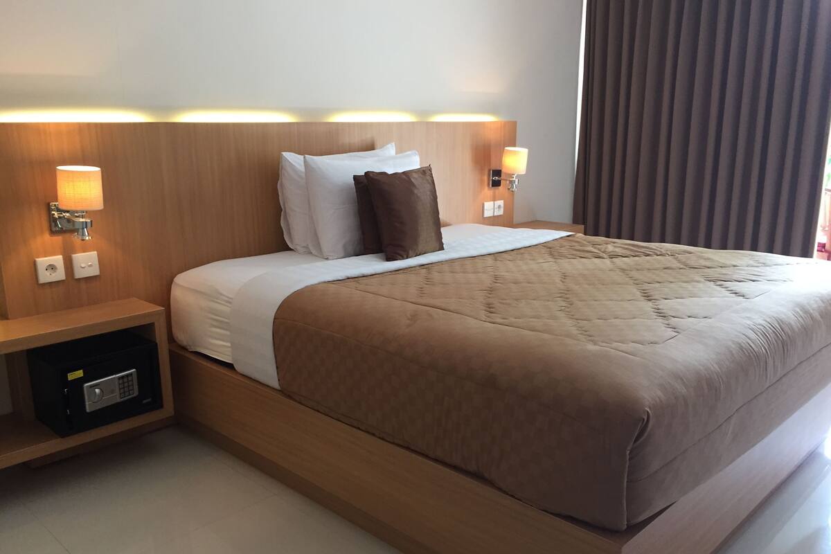 A hotel room with a large bed, wooden headboard, bedside lamps, and a nightstand with a safe.