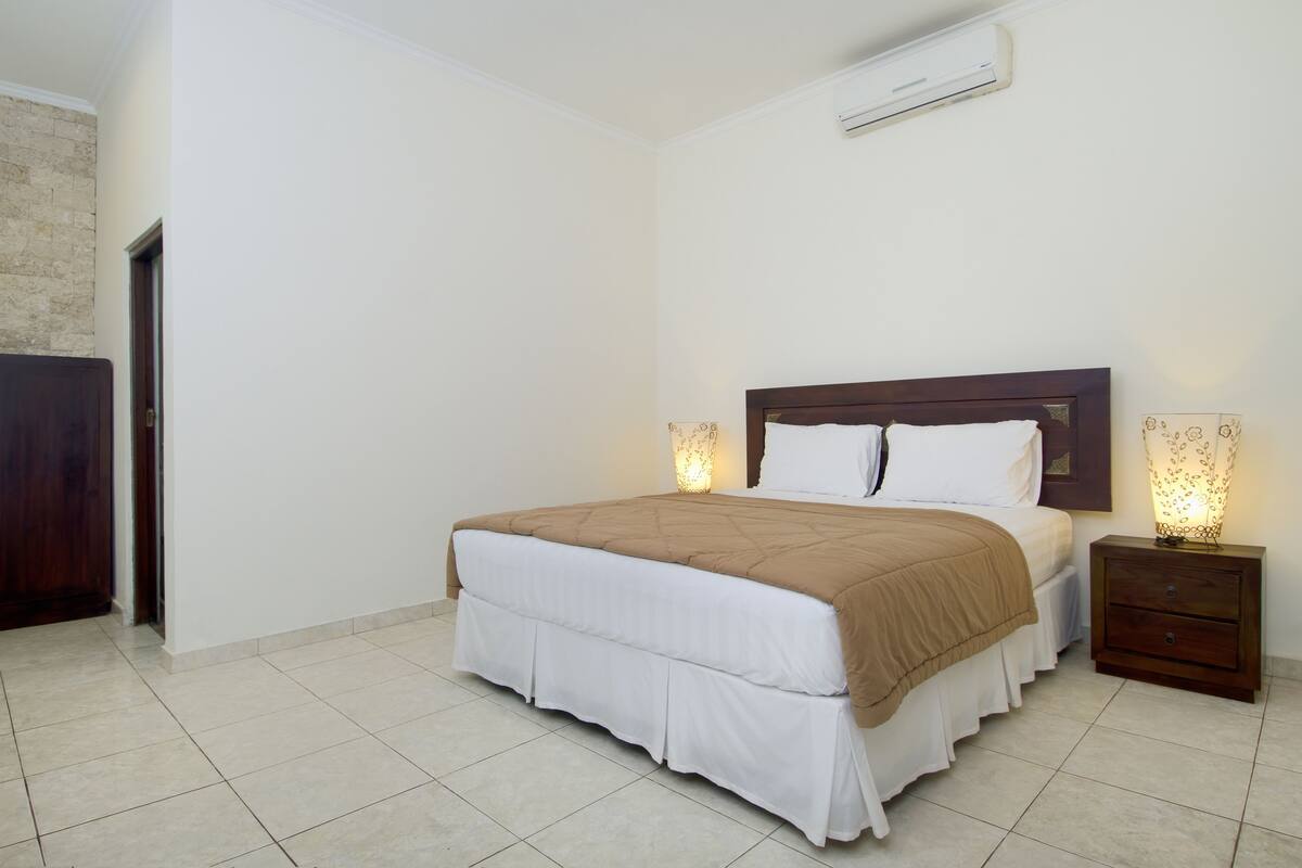 A neatly arranged bedroom with a large bed, a bedside table with a lamp, and a wall-mounted air conditioner.