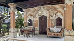 Un área de estar al aire libre con una mesa y sillas de madera, un sofá con cojines, y una entrada decorativa con detalles tallados y un colgador de luz.
