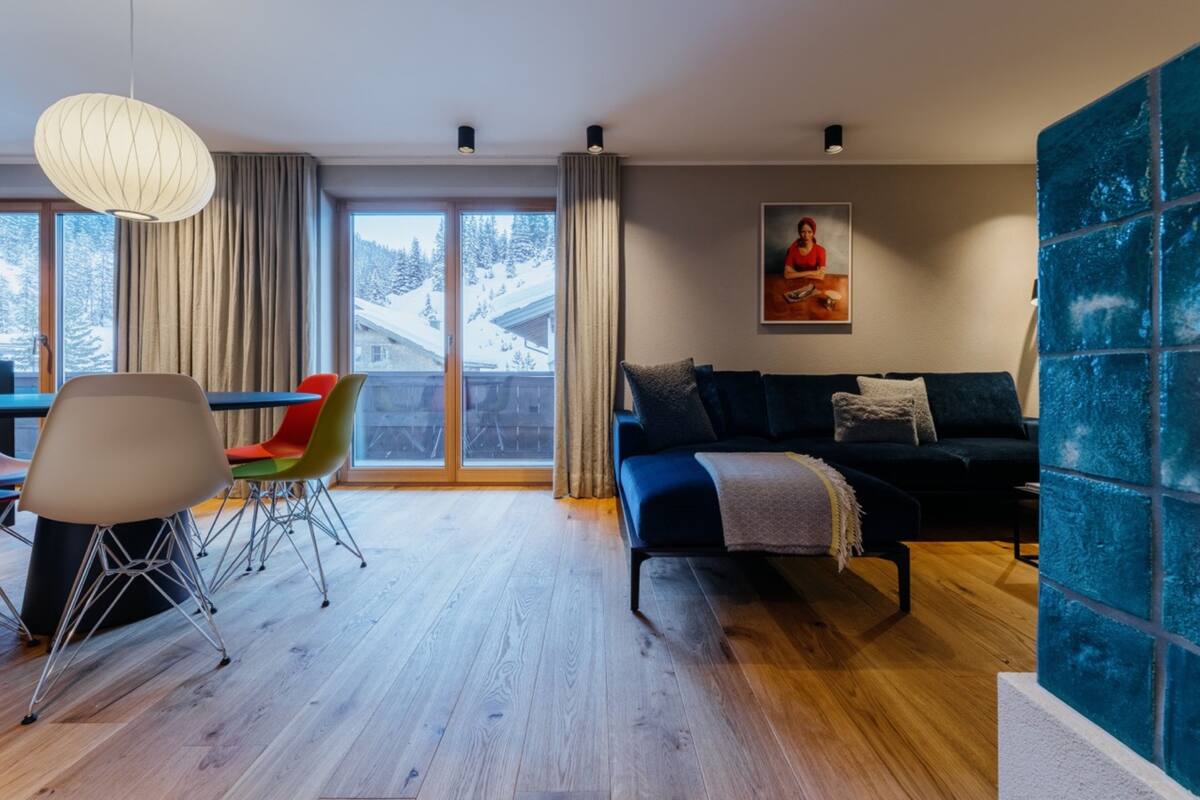 Un soggiorno moderno con un divano, un tavolo da pranzo con sedie colorate e un'ampia finestra vista su montagne innevate.