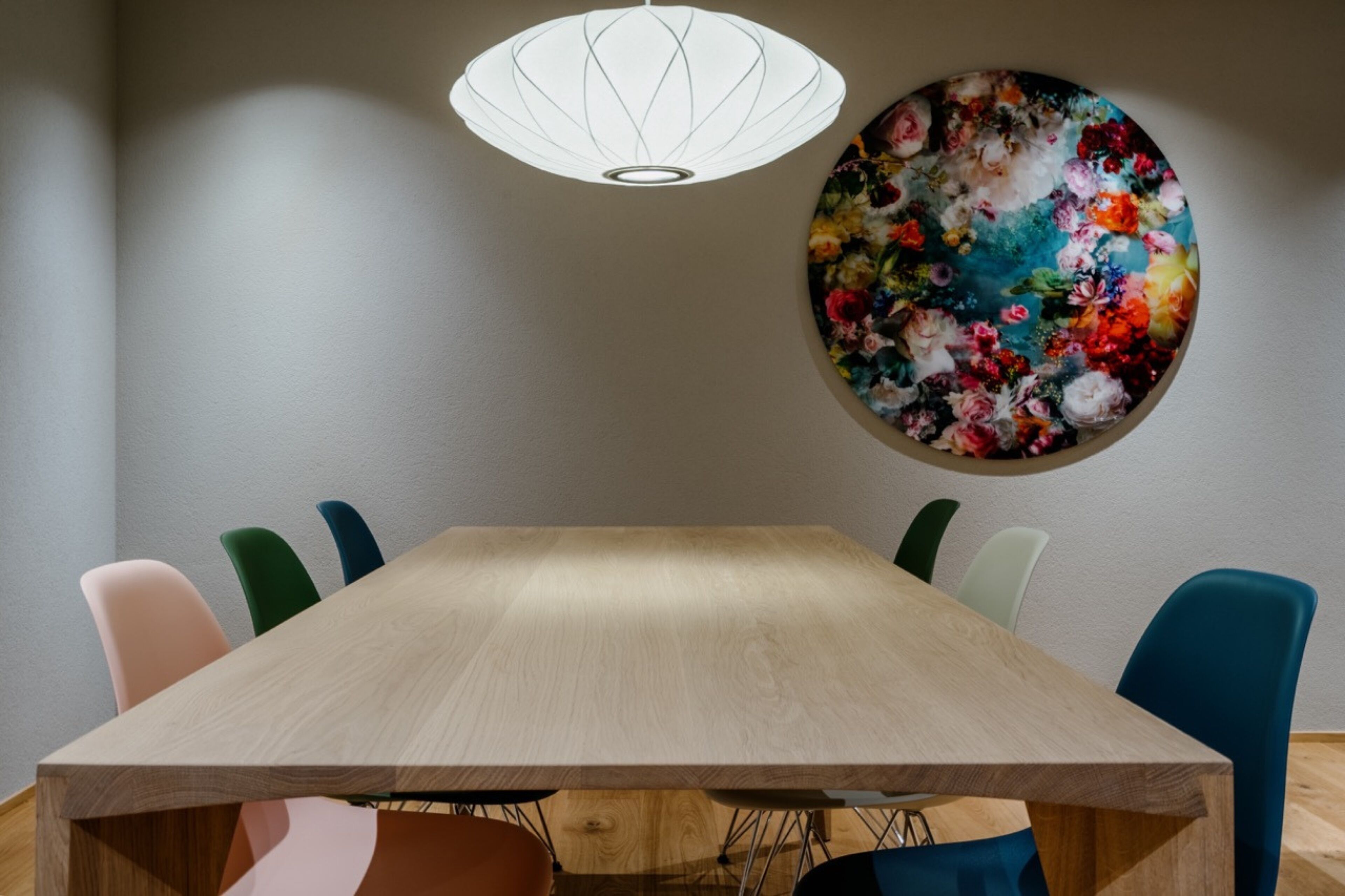 A conference room with a wooden table, colorful chairs, a white pendant light, and a floral painting on the wall.