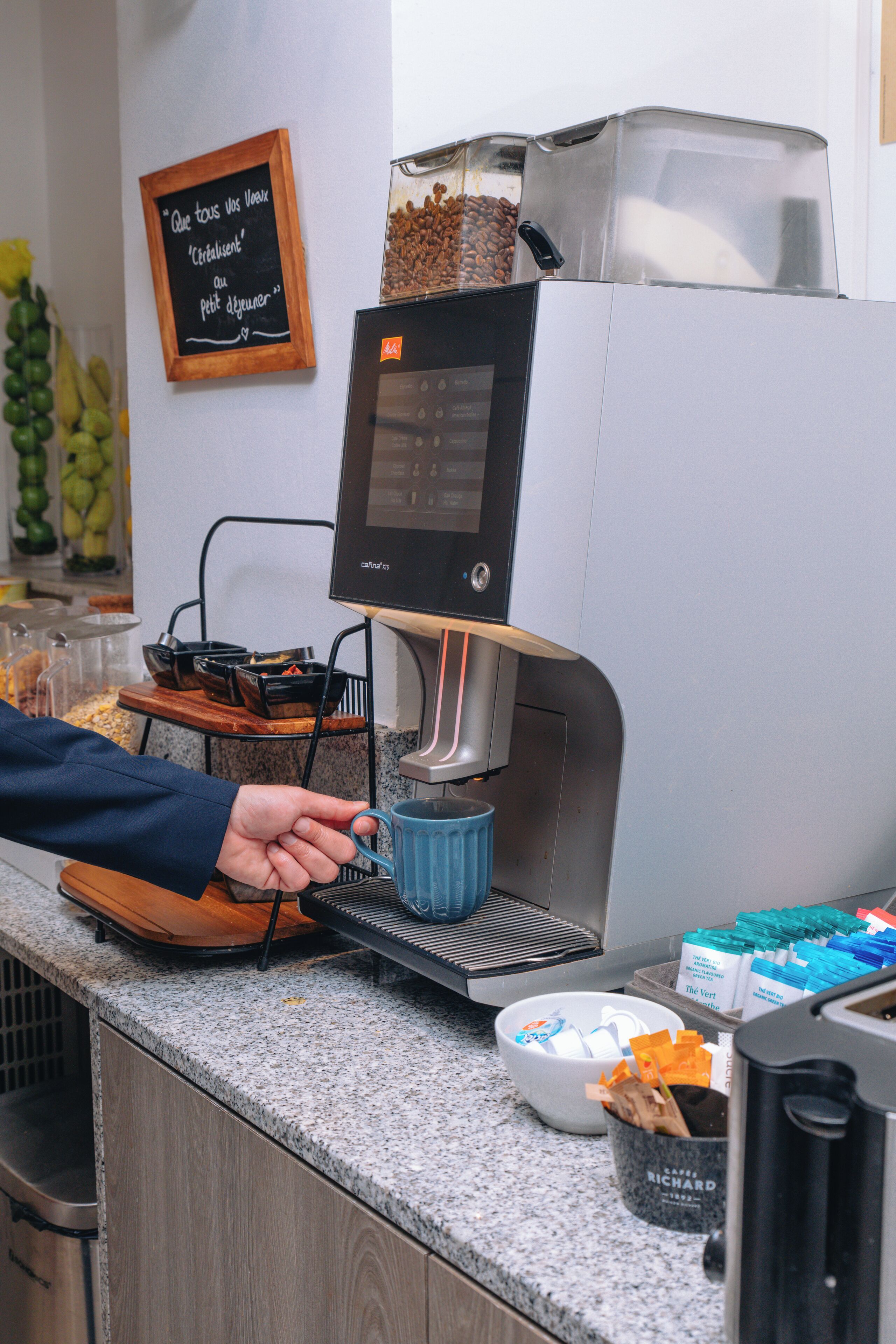 Une machine à café dotée d’un écran numérique, une tasse à café et un plateau contenant des grains de café et un récipient de café moulu.