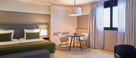A modern hotel room with a bed, a desk with chairs, a window with curtains, and a ceiling light.