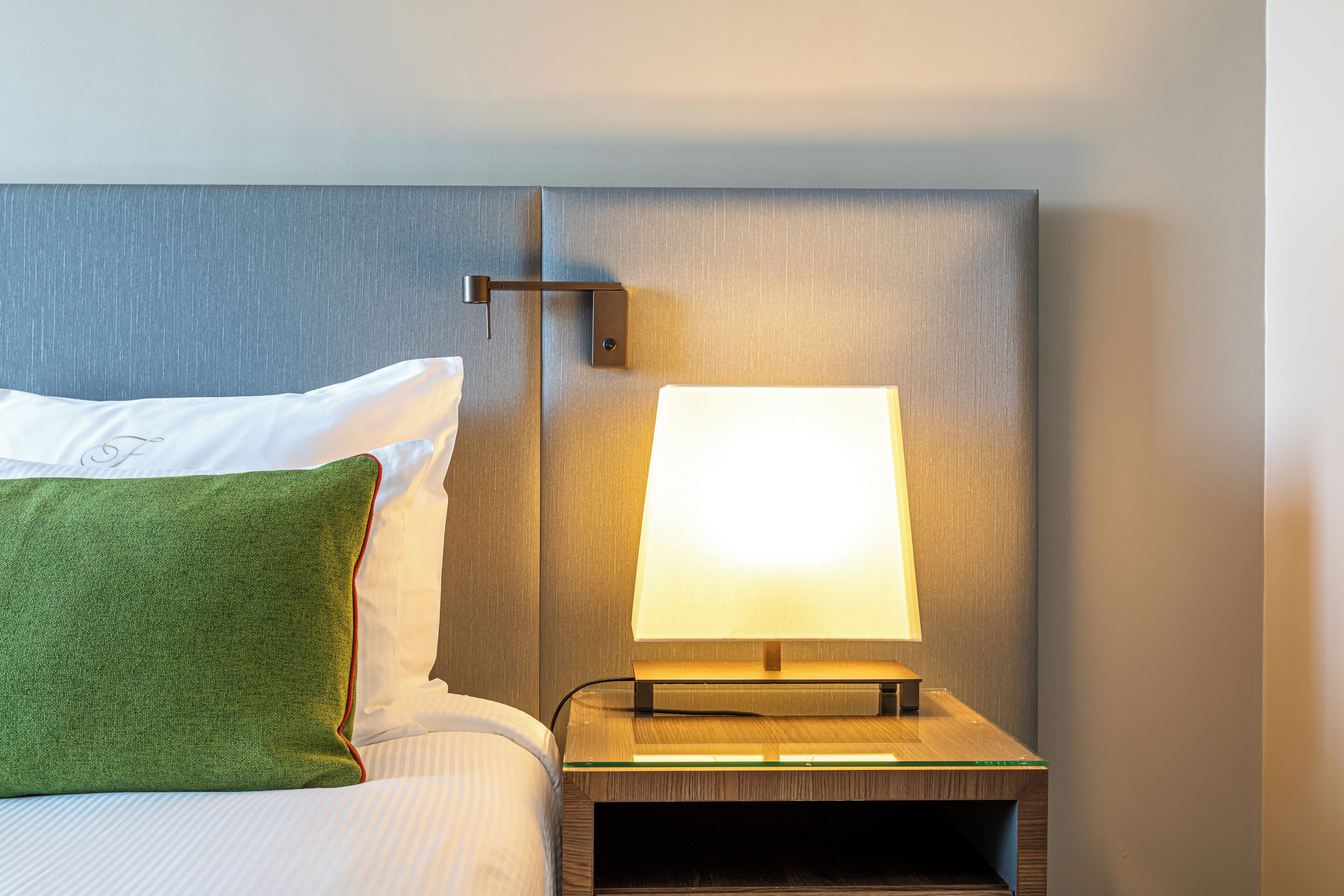 A neatly made bed with white linens and a green pillow, a bedside table with a lamp, and a wall-mounted light fixture.