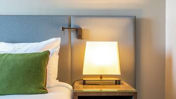 A neatly made bed with white linens and a green pillow, a bedside table with a lamp, and a wall-mounted light fixture.