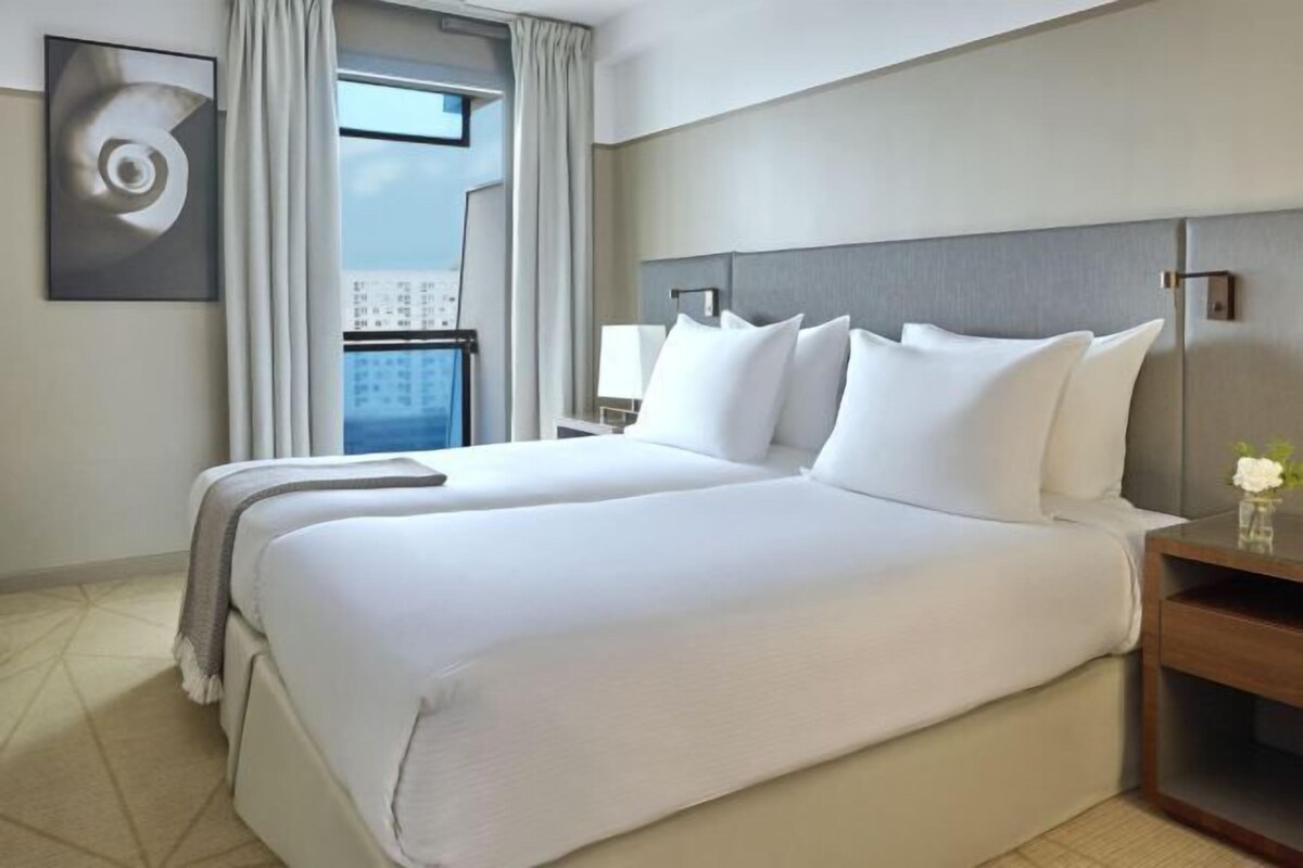 Habitación de hotel con una cama grande, una mesita de noche, una ventana con cortinas y una lámpara fijada a la pared.