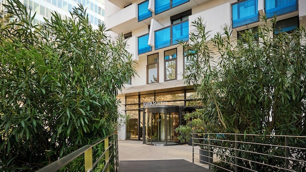 A hotel entrance with a revolving door, surrounded by lush greenery and a modern building facade with blue-framed windows.