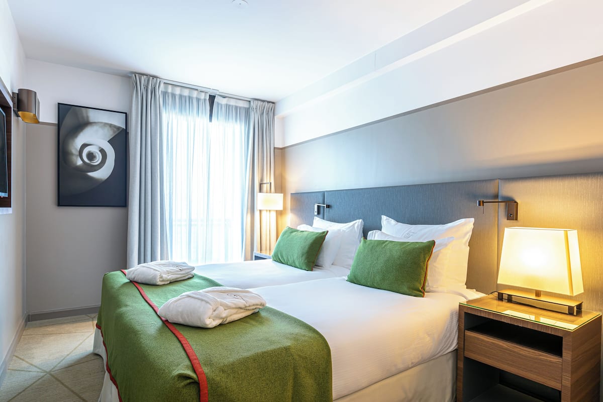 Ein Hotelzimmer mit zwei Betten, einem großen Fenster mit Vorhängen, einem Wandbild und einer Nachttischlampe.