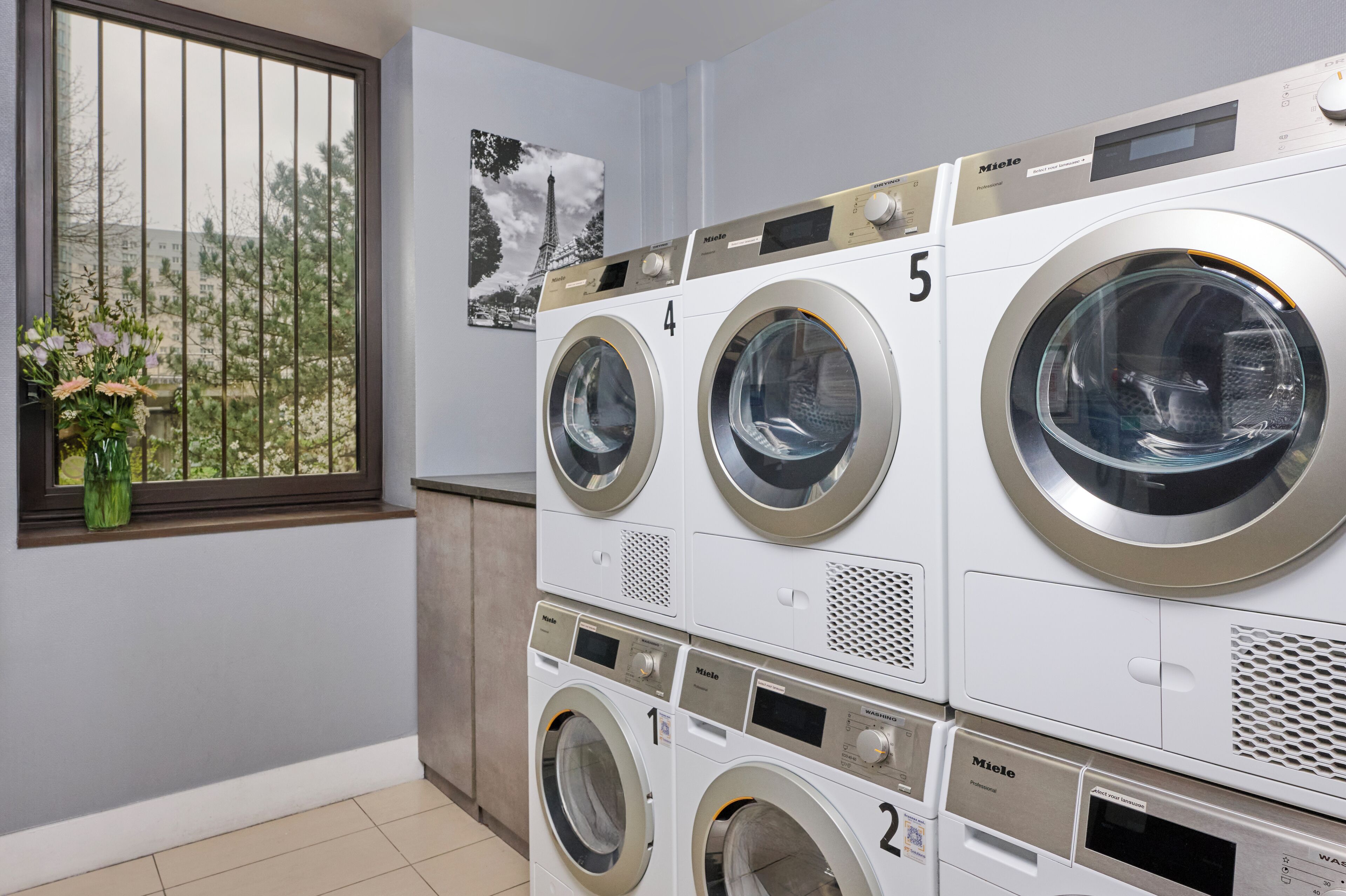 A laundry room with five stacked front-loading washing machines, a window with a view of trees, and a picture of the Eiffel Tower on the wall.