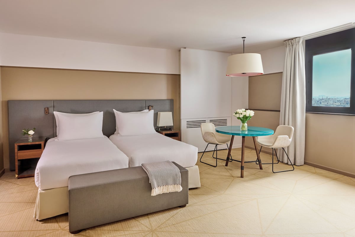 Ein Hotelzimmer mit Doppelbett, kleinem Tisch mit zwei Stühlen, einer Bank, einem Nachttisch und einem Fenster mit Blick auf die Stadt.
