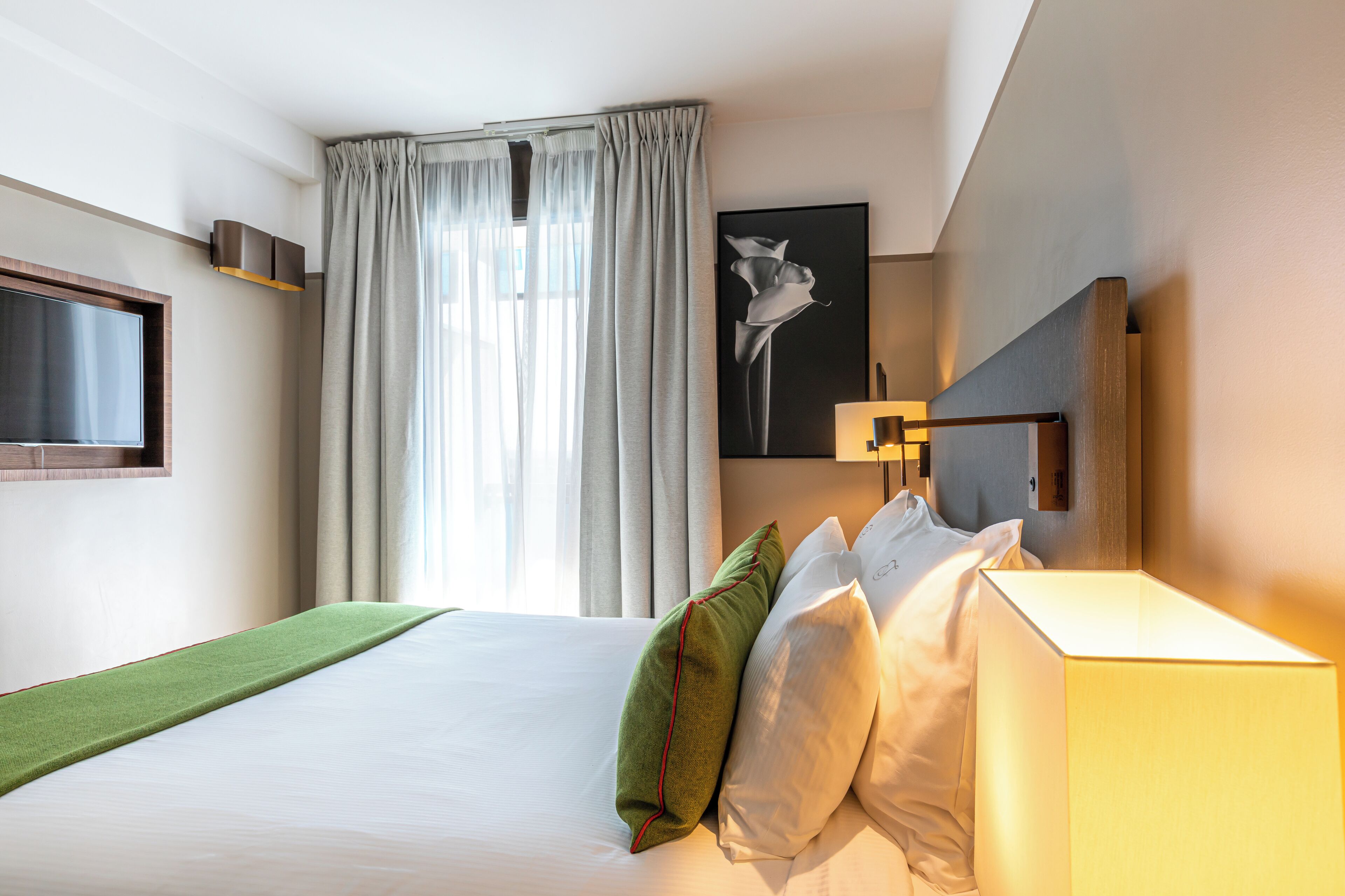 Une chambre d’hôtel avec un lit, un téléviseur à écran plat, un tableau, une lampe et une fenêtre avec des rideaux.
