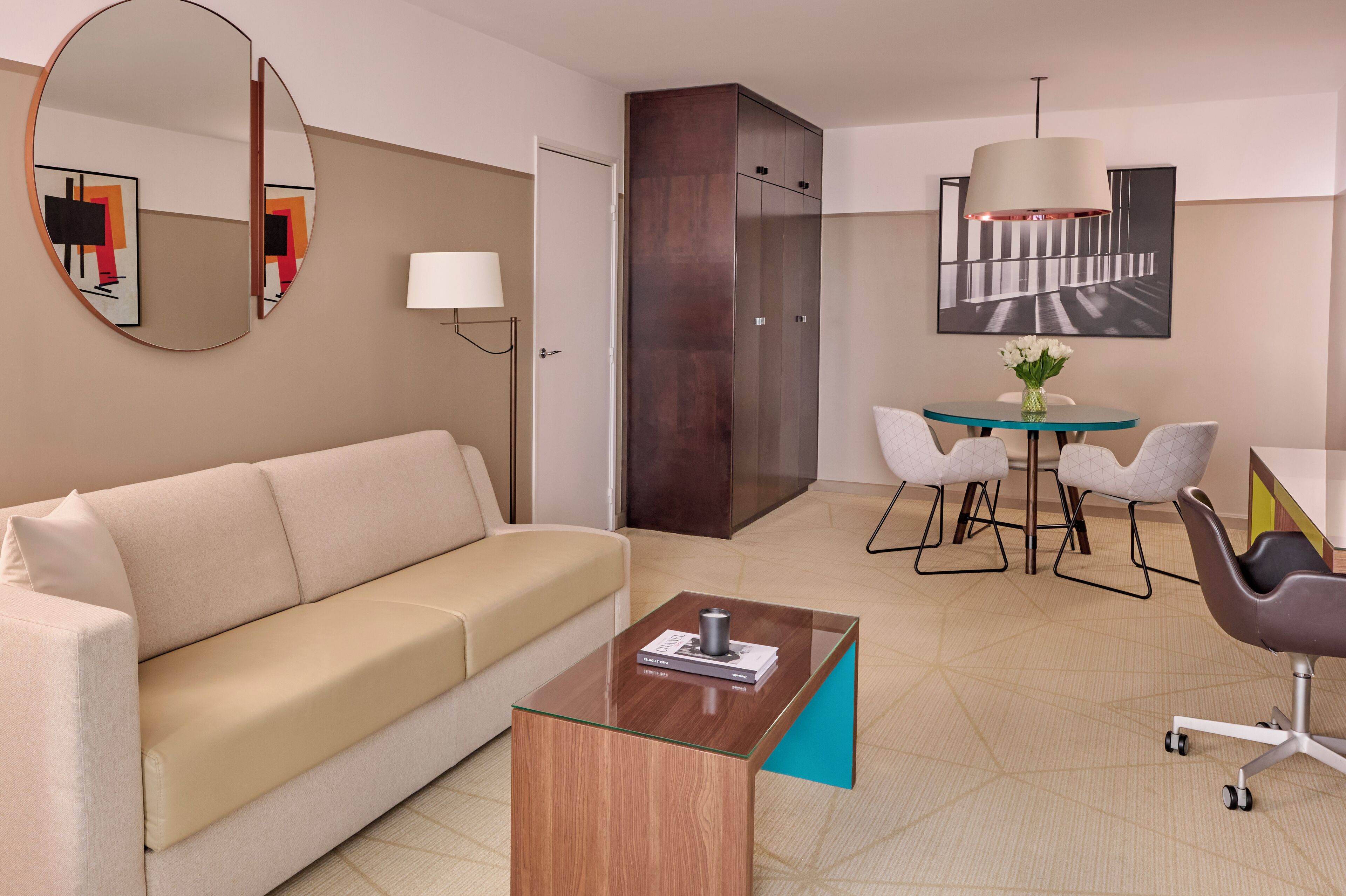 Una habitación de hotel moderna con sofá, una mesita de centro, zona de comedor con sillas, un espejo grande y un cuadro en la pared.