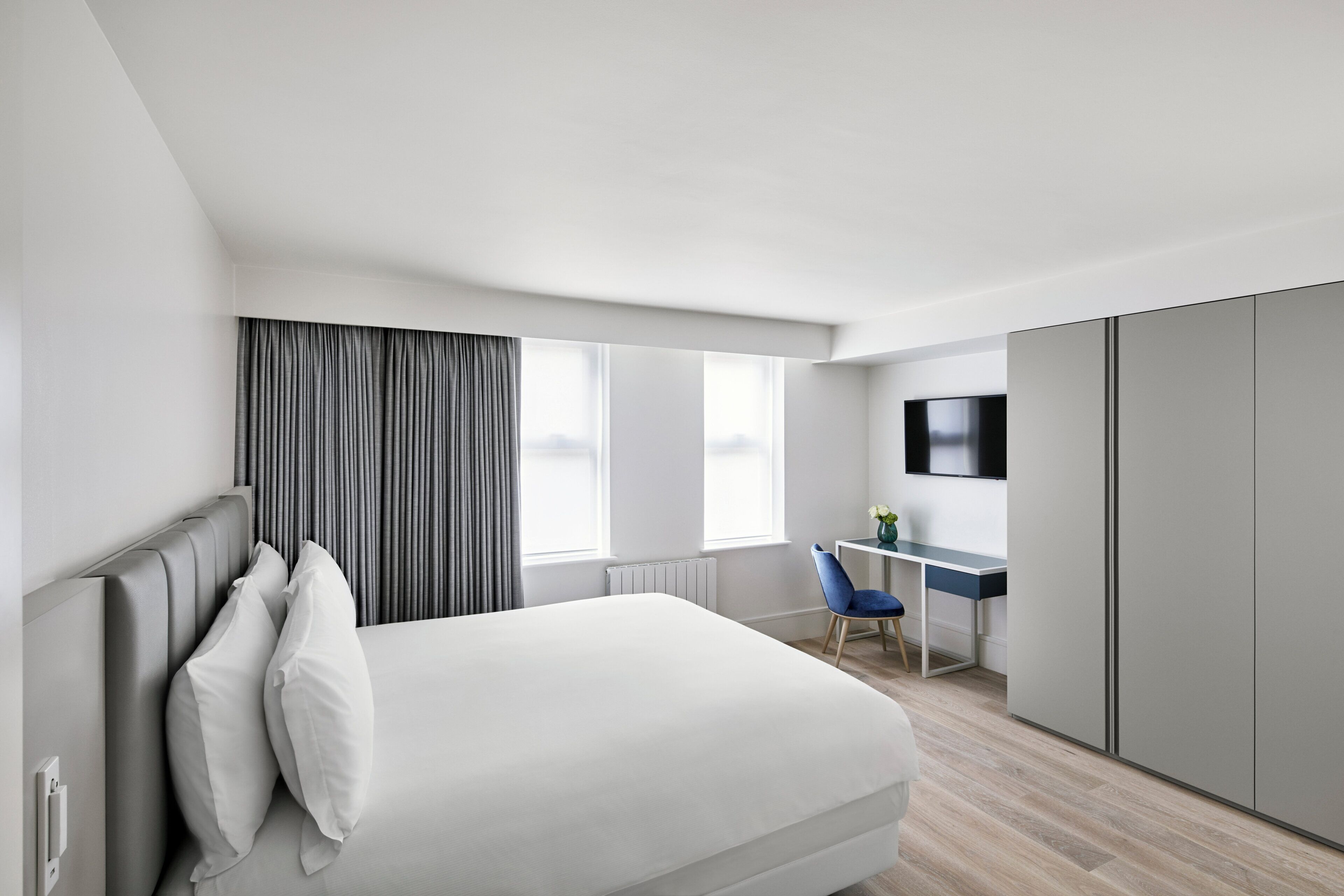 A modern hotel room with a bed, a desk, a chair, a TV, and a wardrobe.