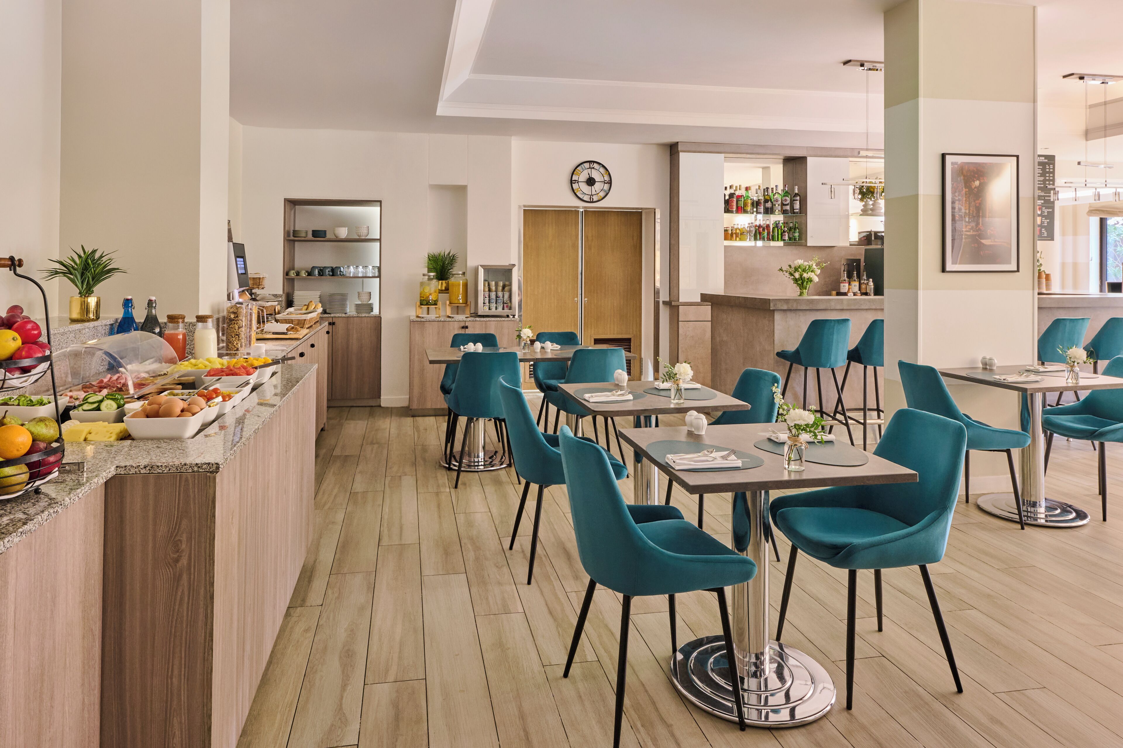 A modern dining area with teal chairs, a wooden counter with fruits and beverages, and a bar area with bottles and glasses.