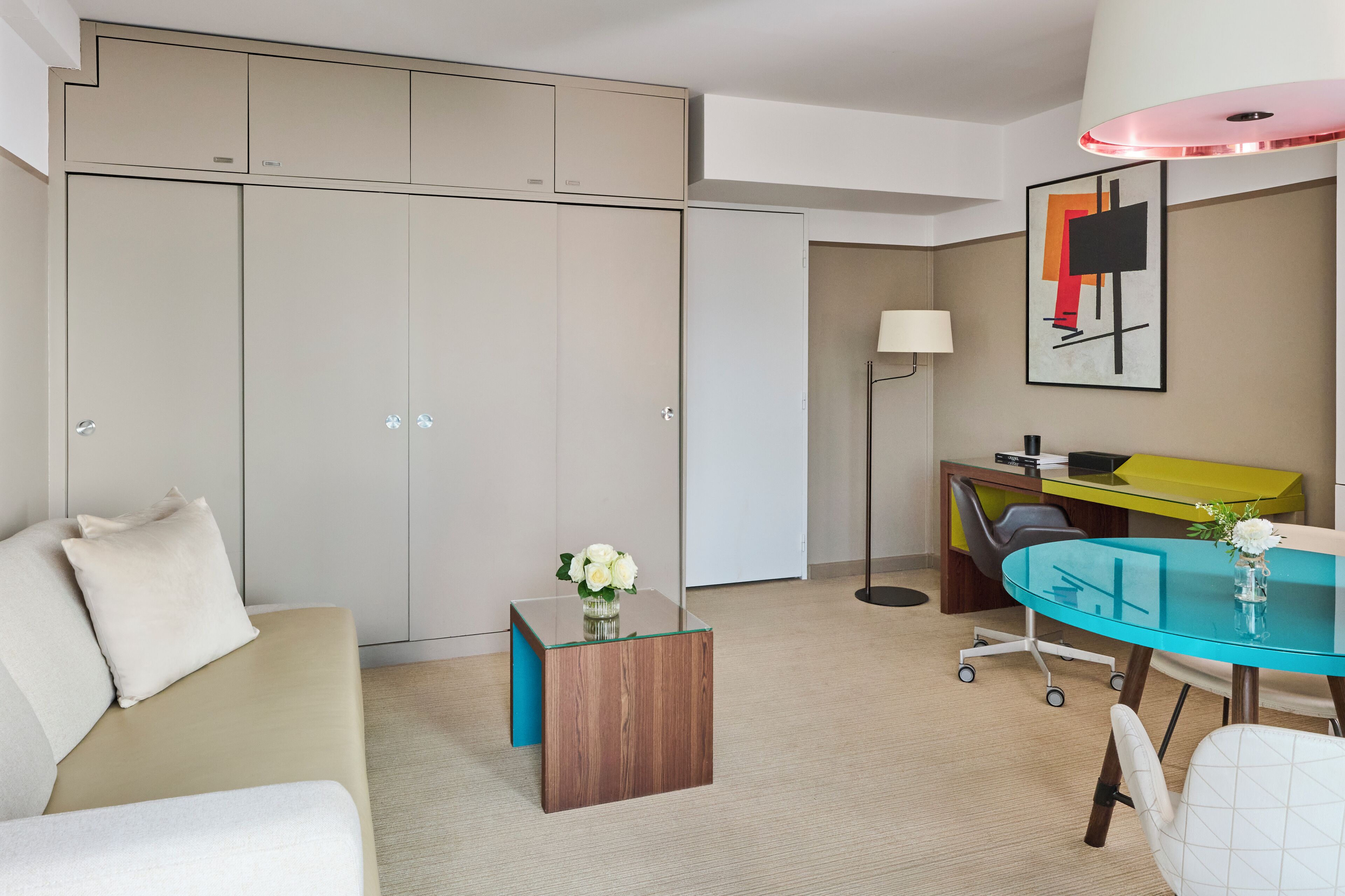 A modern hotel room with a sofa, a glass coffee table, a desk with a chair, a wardrobe, and a lamp.