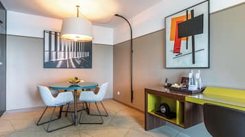 Une chambre d’hôtel moderne dotée d’un coin repas comprenant une table ronde et deux chaises, un bureau avec une chaise, une lampe et un tableau abstrait encadré accroché au mur.