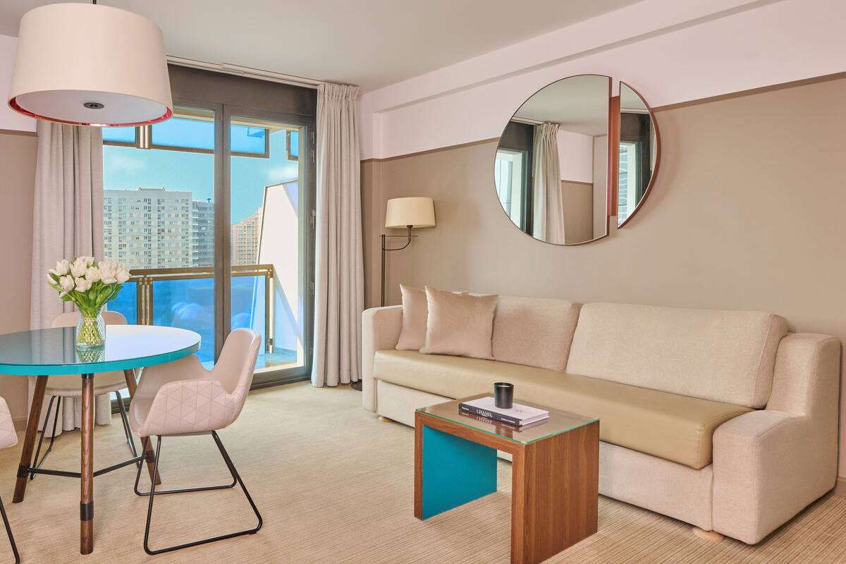 Ein modernes Hotelzimmer mit einem runden Spiegel, einem beigen Sofa, einem Glastisch und Blick auf die Stadt durch große Fenster.