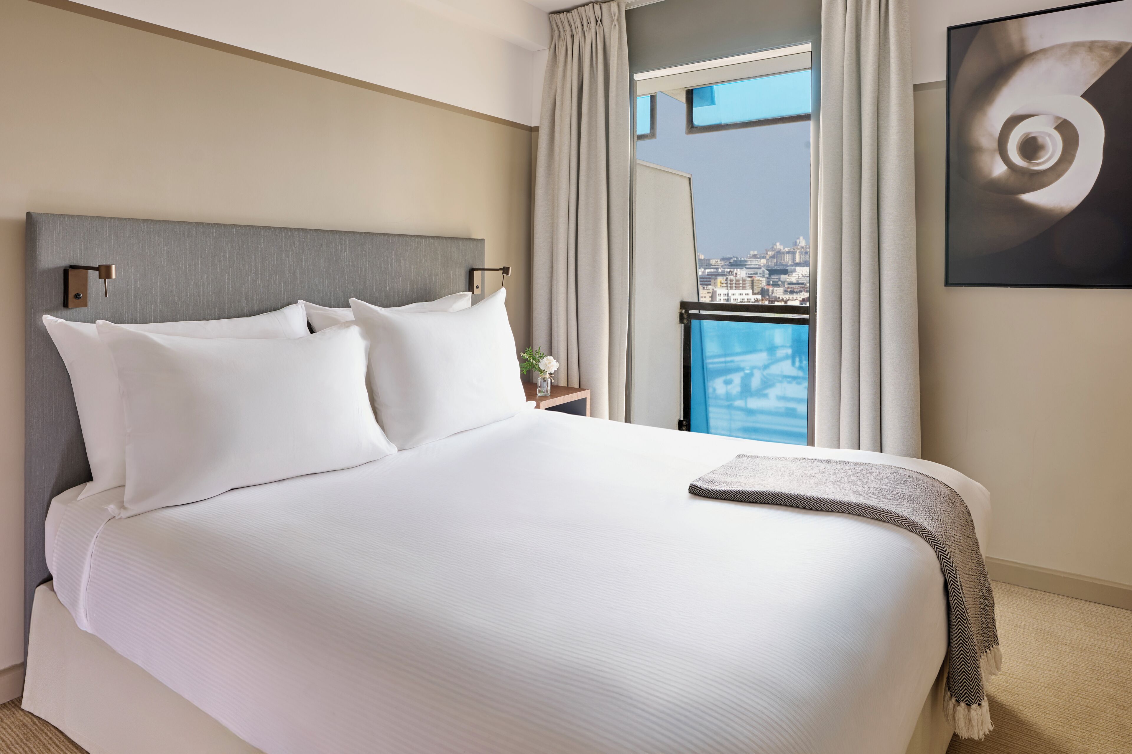 A neatly made bed with white linens, a grey headboard, and a view of a cityscape through a window with curtains.