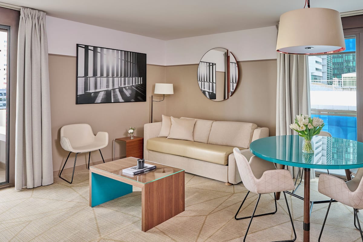 Ein modernes Hotelzimmer mit einer Couch, einem Glastisch, einem runden Esstisch und einem großen Fenster mit Vorhängen.