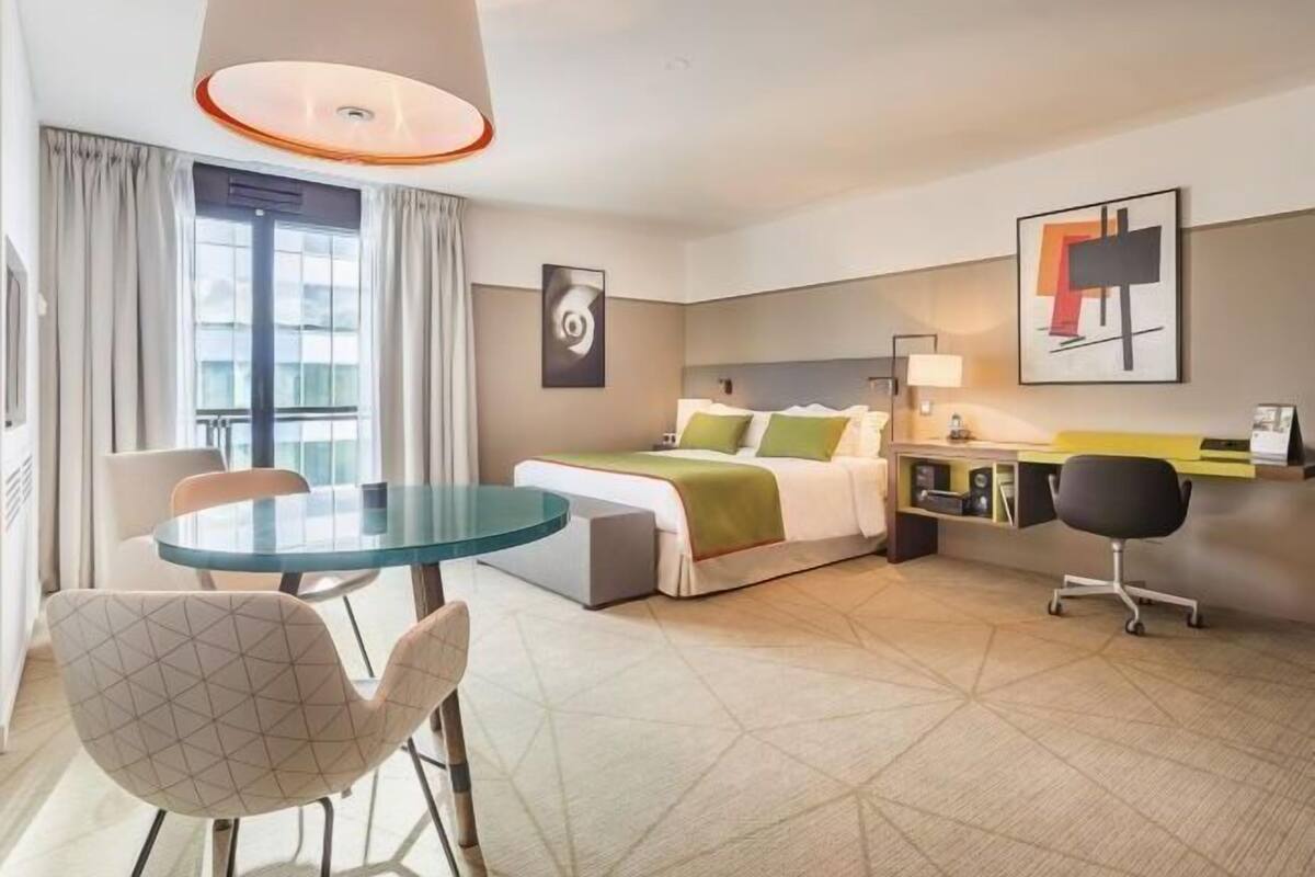 Ein Hotelzimmer mit Bett, Schreibtisch, Stuhl, rundem Tisch und einem Fenster mit Vorhängen.