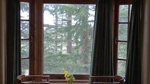 A room with a large window, two wooden chairs, a small table, and a vase with flowers. - The Woodstock Resort (Shimla)