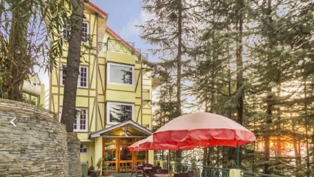 An outdoor patio with tables and chairs, a stone wall, and a building with a yellow facade and a red roof. - The Woodstock Resort (Shimla)