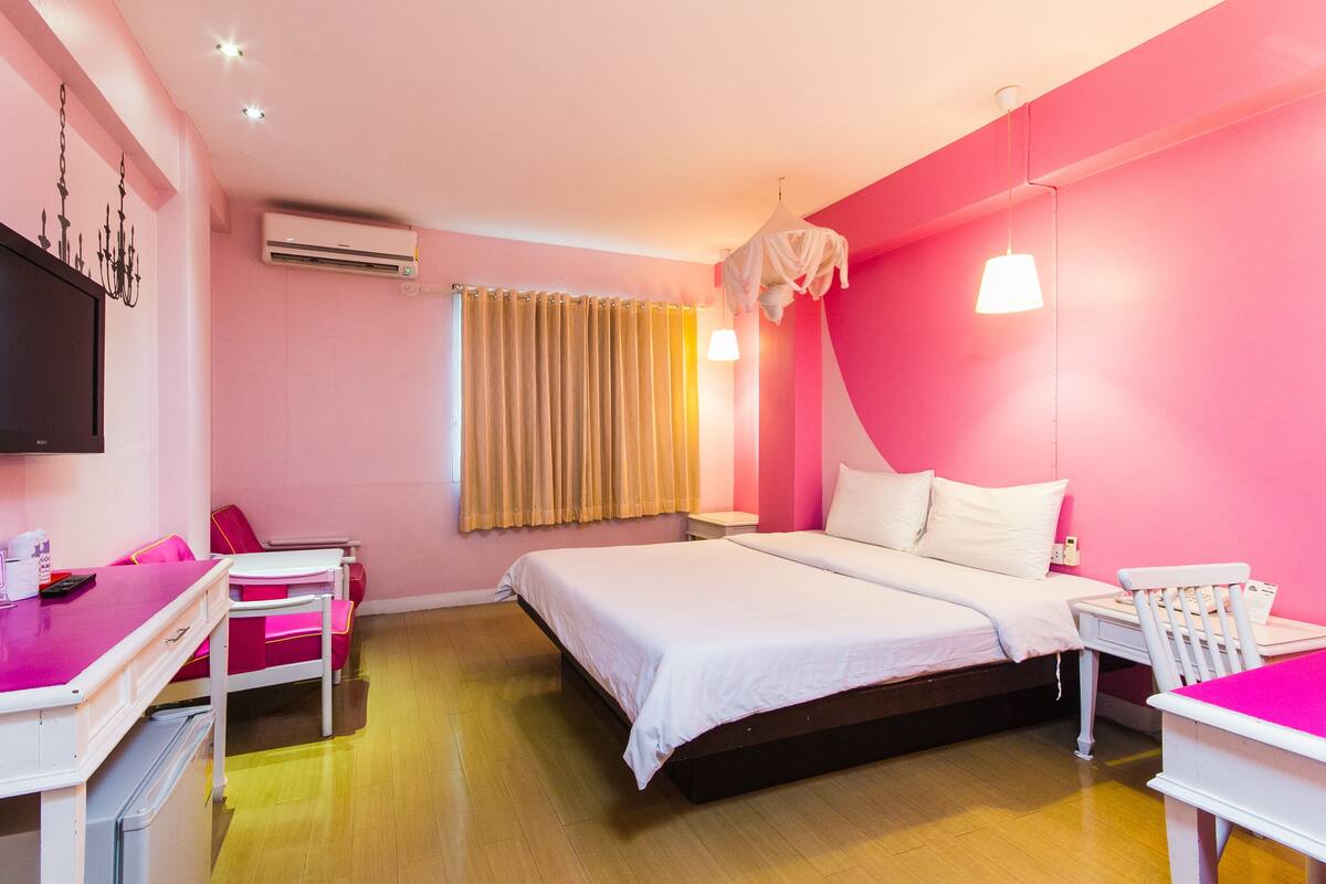 A hotel room with a bed, bedside tables, a desk, a chair, a TV, an air conditioner, and a window with curtains.