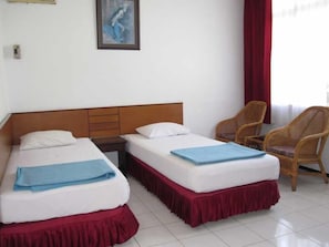 A hotel room with two beds, a wooden headboard, a red skirted bed, a blue blanket, a wicker chair, and a painting on the wall. - Hotel Tosari (Malang)