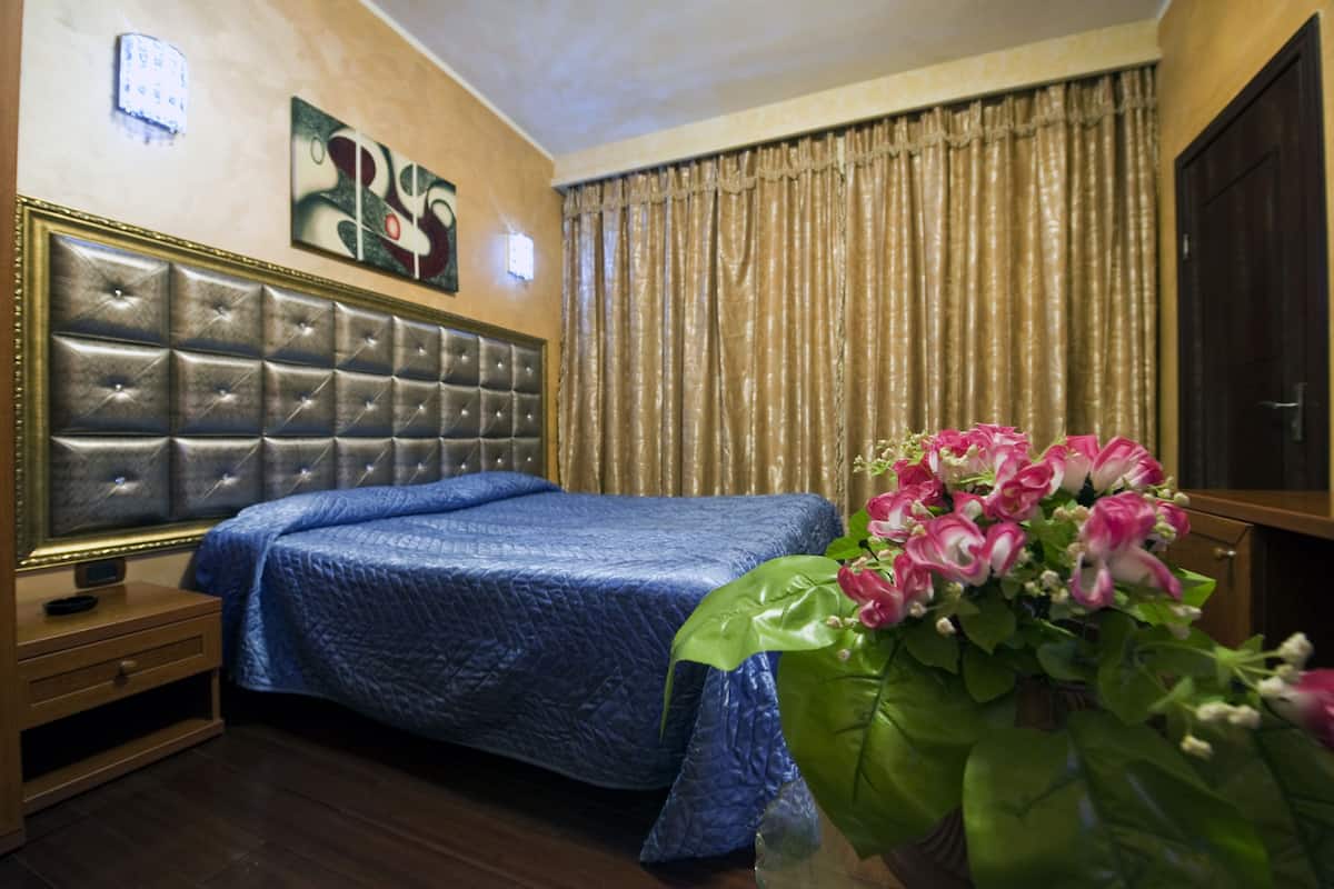 A hotel room with a large bed, a wooden nightstand, a bedside table, a wall-mounted TV, and a window with curtains.