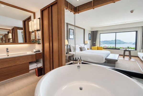 A modern hotel room with a large bathtub, a separate bedroom with a bed and a view, a bathroom with a vanity and mirror, and a large window offering a scenic view. - The Nai Harn (Rawai)