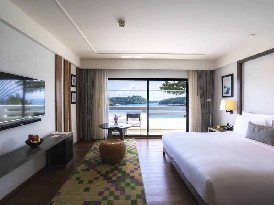 A hotel room with a large bed, a TV, a desk with a bowl of fruit, a chair, a small table with a vase, and a view of a body of water through large windows.