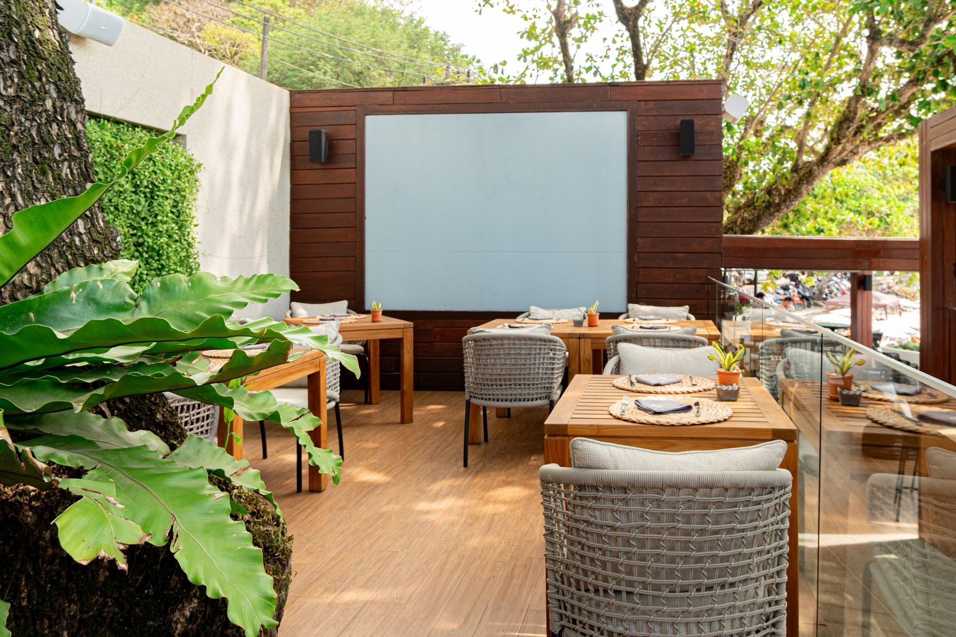 An outdoor dining area with wooden tables and wicker chairs, a large screen, and a glass railing overlooking a garden.