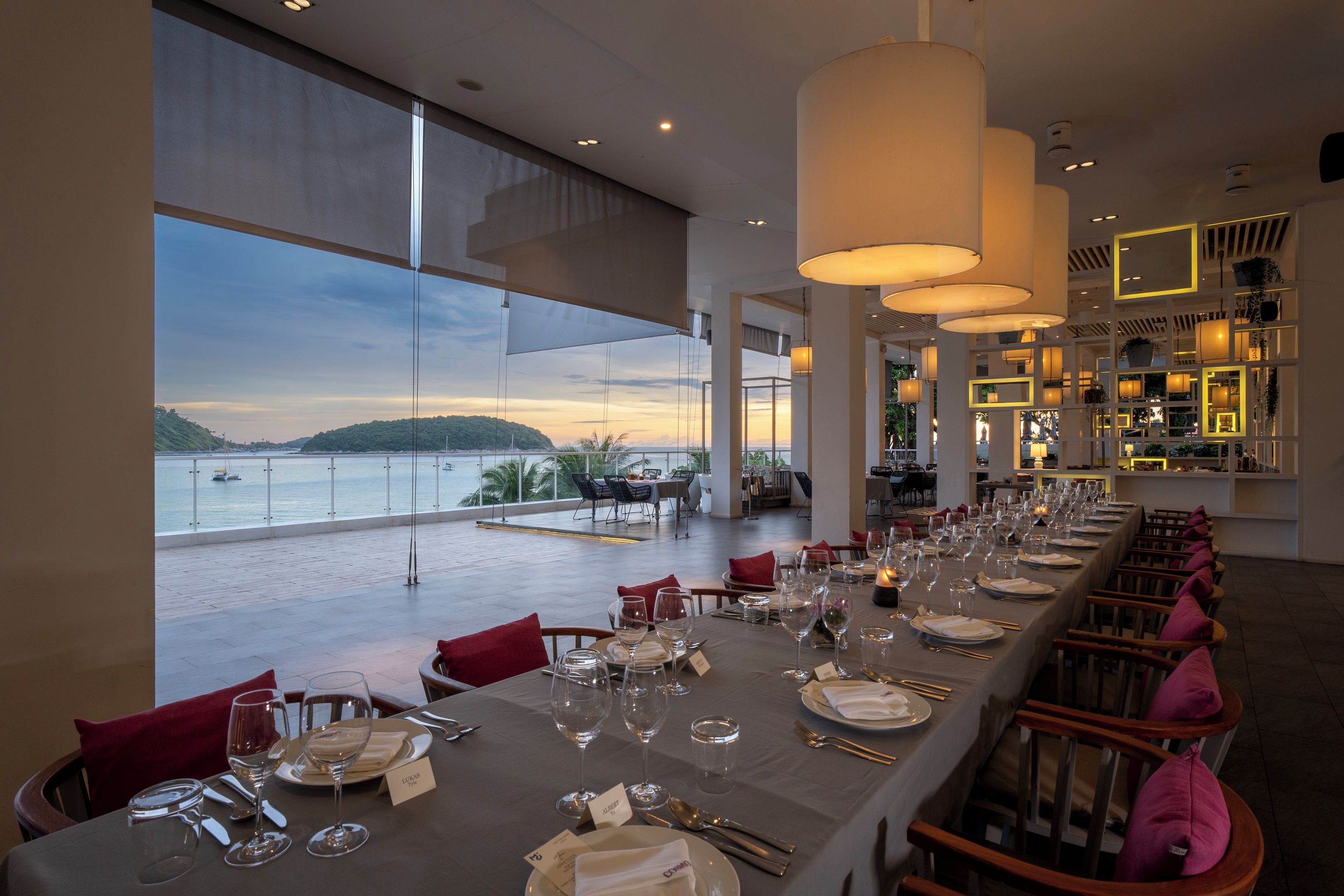 A restaurant with a long dining table set for a meal, overlooking a scenic view of the sea and hills.