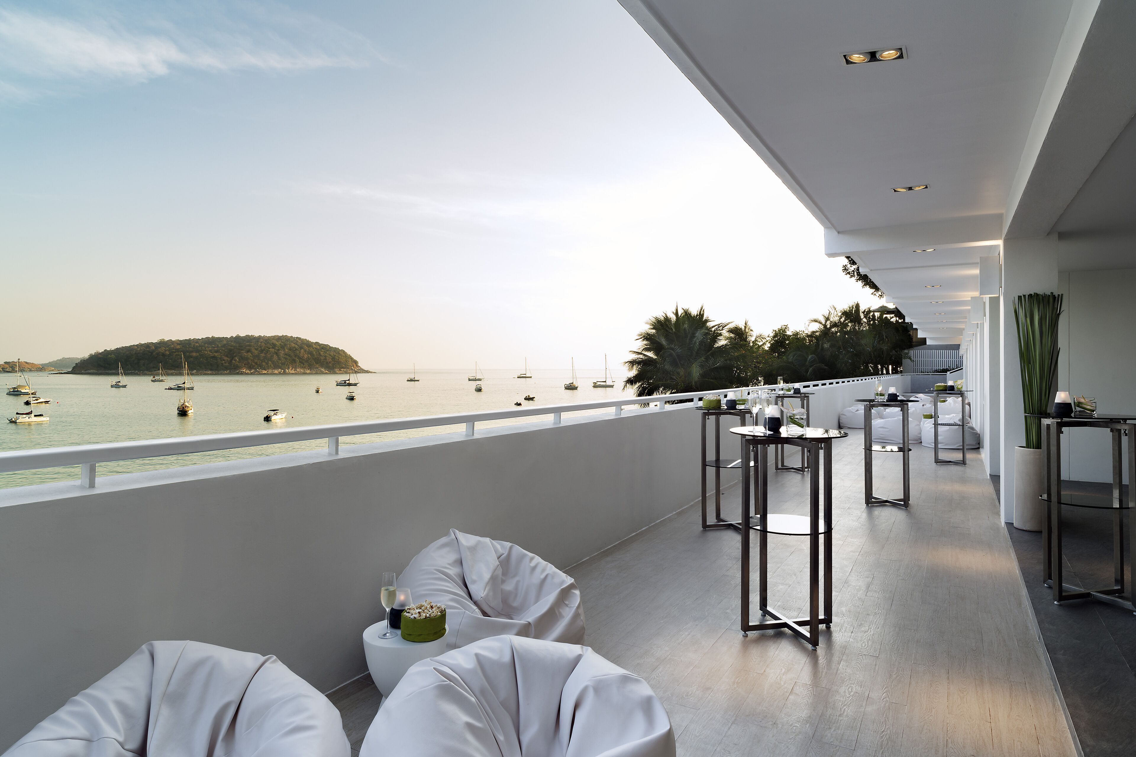 A balcony with white cushioned seating, a small table with a candle, and a view of the sea with boats.