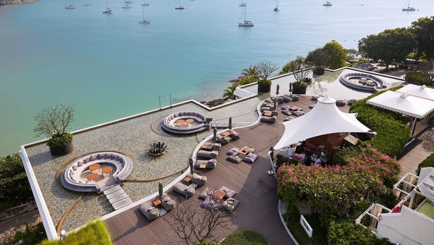A coastal resort with a circular outdoor seating area, a covered patio, and a view of the sea with sailboats. - The Nai Harn (Rawai)