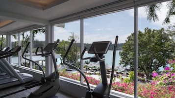 Un gimnasio con cintas de correr y equipo de ejercicio, ventanas amplias con vistas a una playa y palmeras.