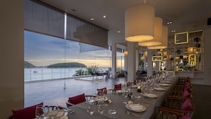 A dining area with a view of the sea, tables set with wine glasses and plates, and modern lighting fixtures. - The Nai Harn (Rawai)