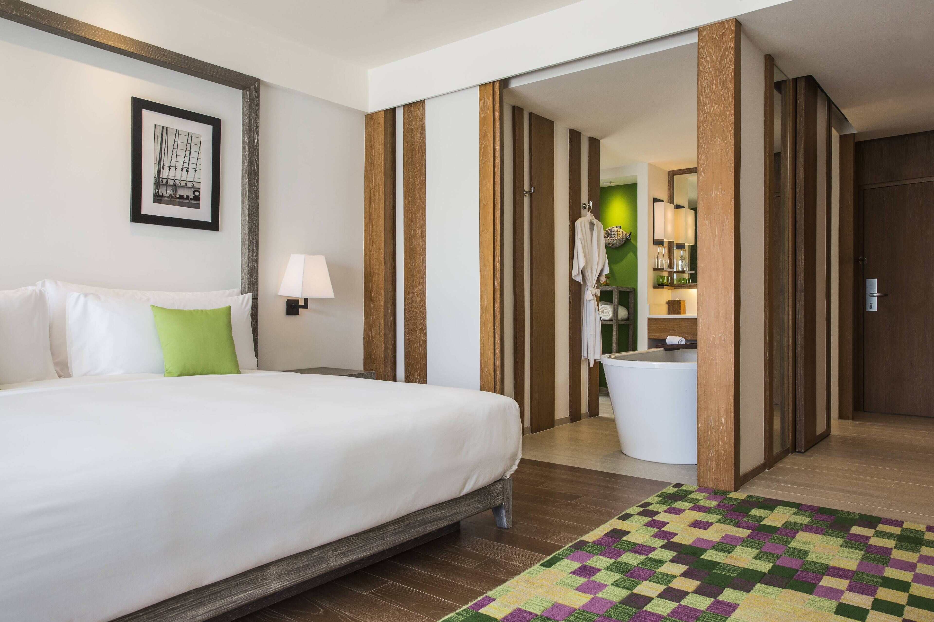 Una habitación de hotel con una cama, un almohadón verde, un tapete de cuadros, suelo de madera, un cuadro enmarcado en la pared y un baño con una bañera blanca y un perchero.