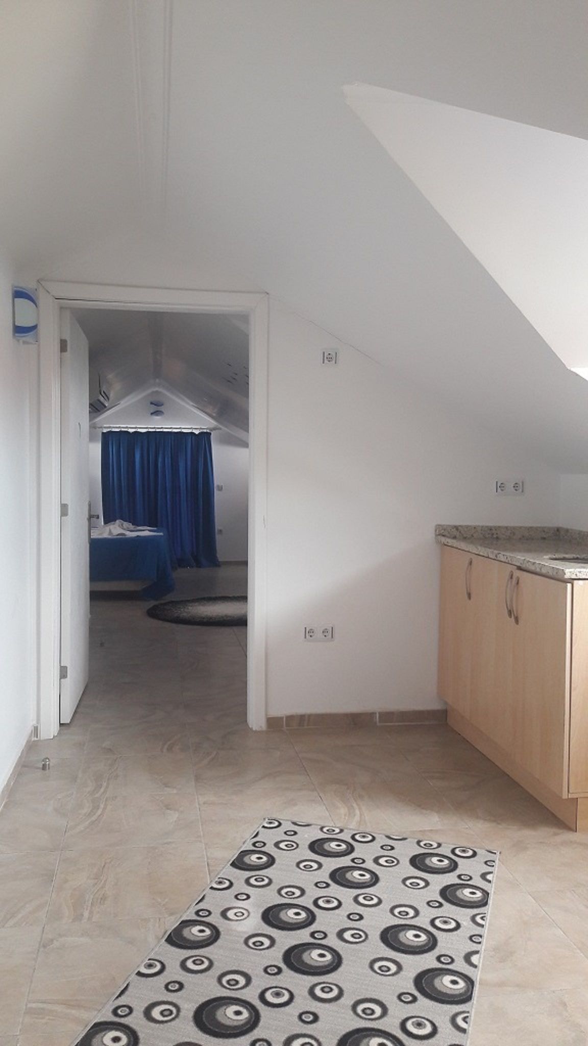A narrow hallway with a patterned rug, a kitchenette with wooden cabinets, and a bedroom with a bed and blue curtains.