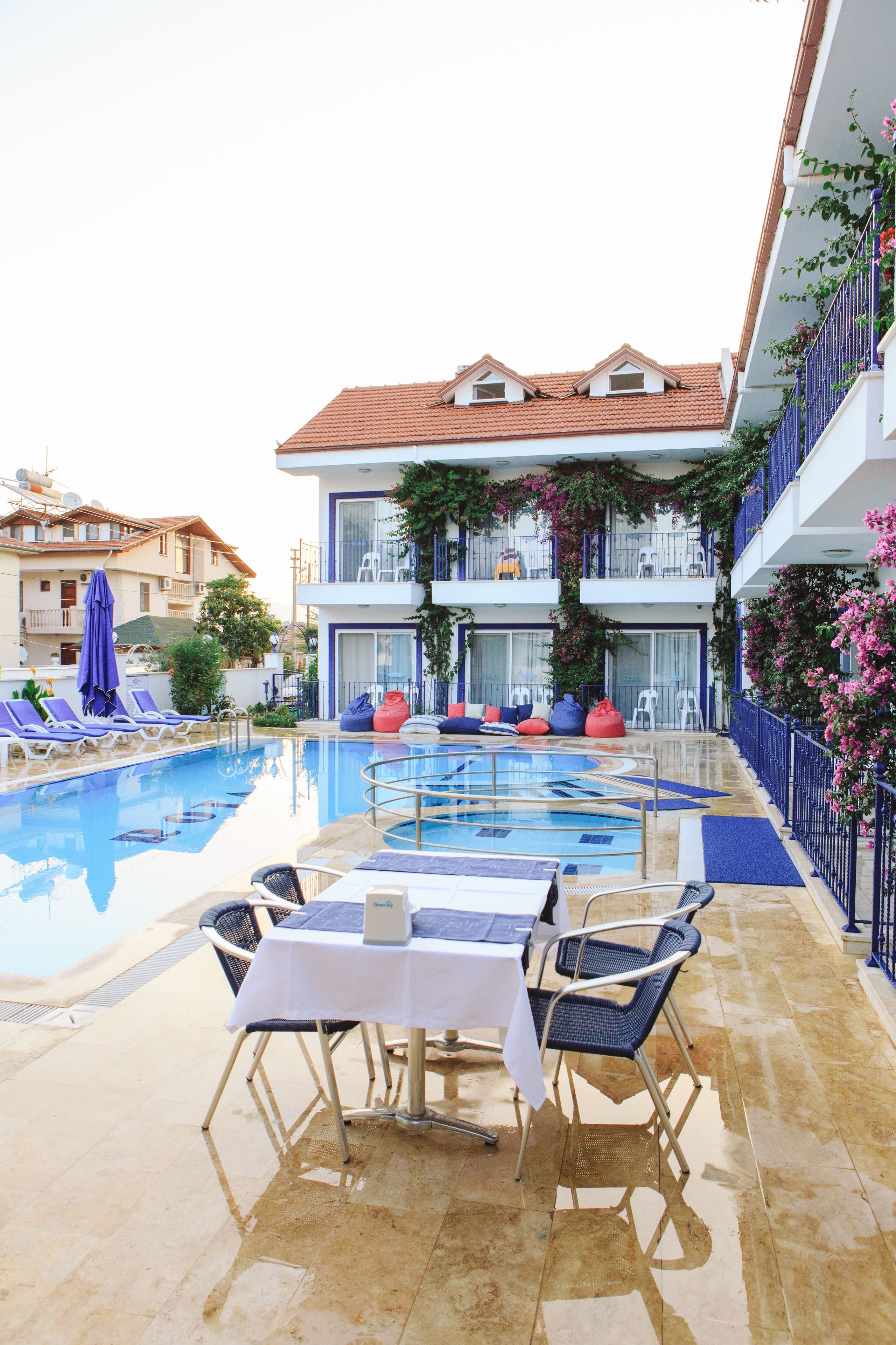 Zona de piscina con sillones reclinables, una mesa y vistas a un edificio con balcones.