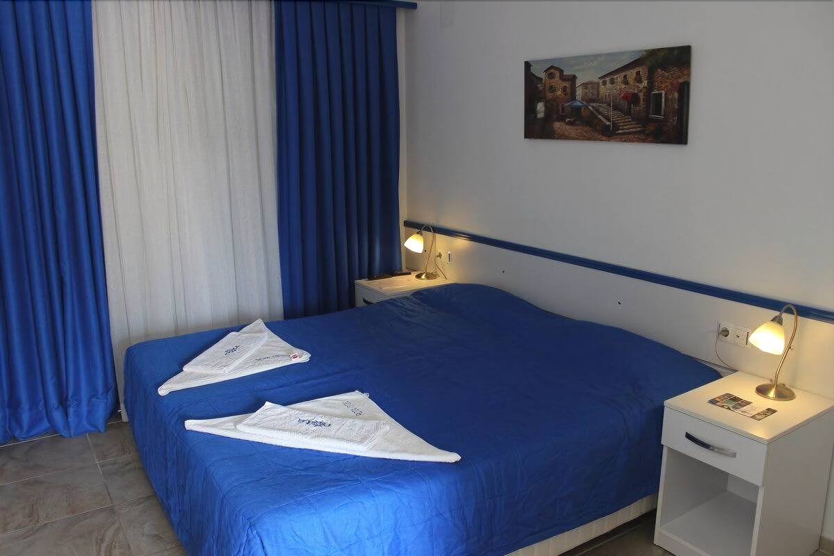 Hotellihuone, jossa on sänky, kaksi yöpöytävalaisinta, yöpöytä ja taulu seinällä.